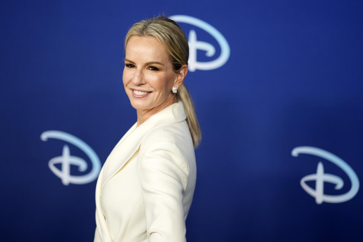 Dr. Jennifer Ashton smiling in a cream-colored suit and low ponytail while standing sideways in front of a blue background