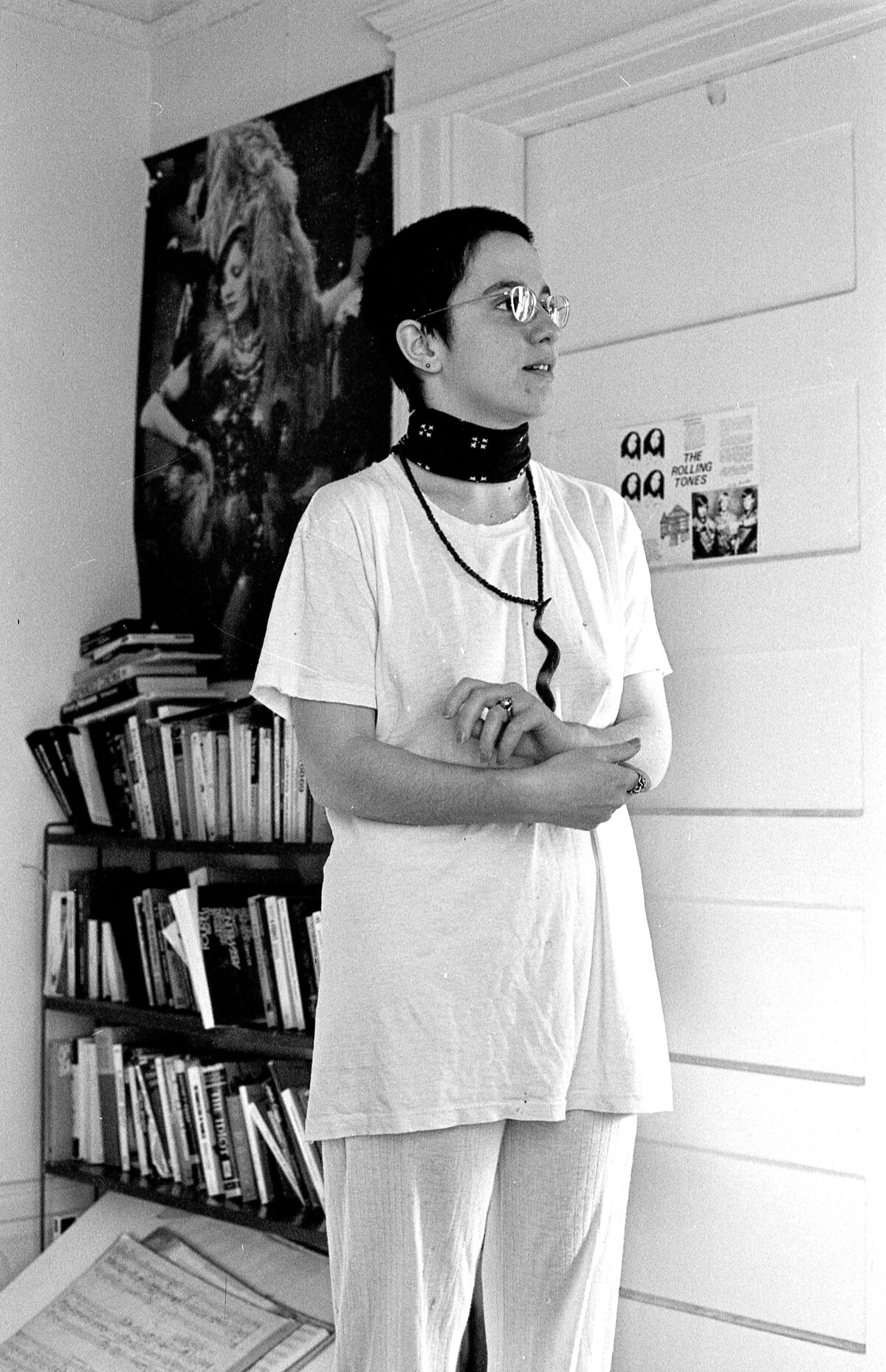 A woman with a buzz cut in an apartment with a bookshelf behind her 