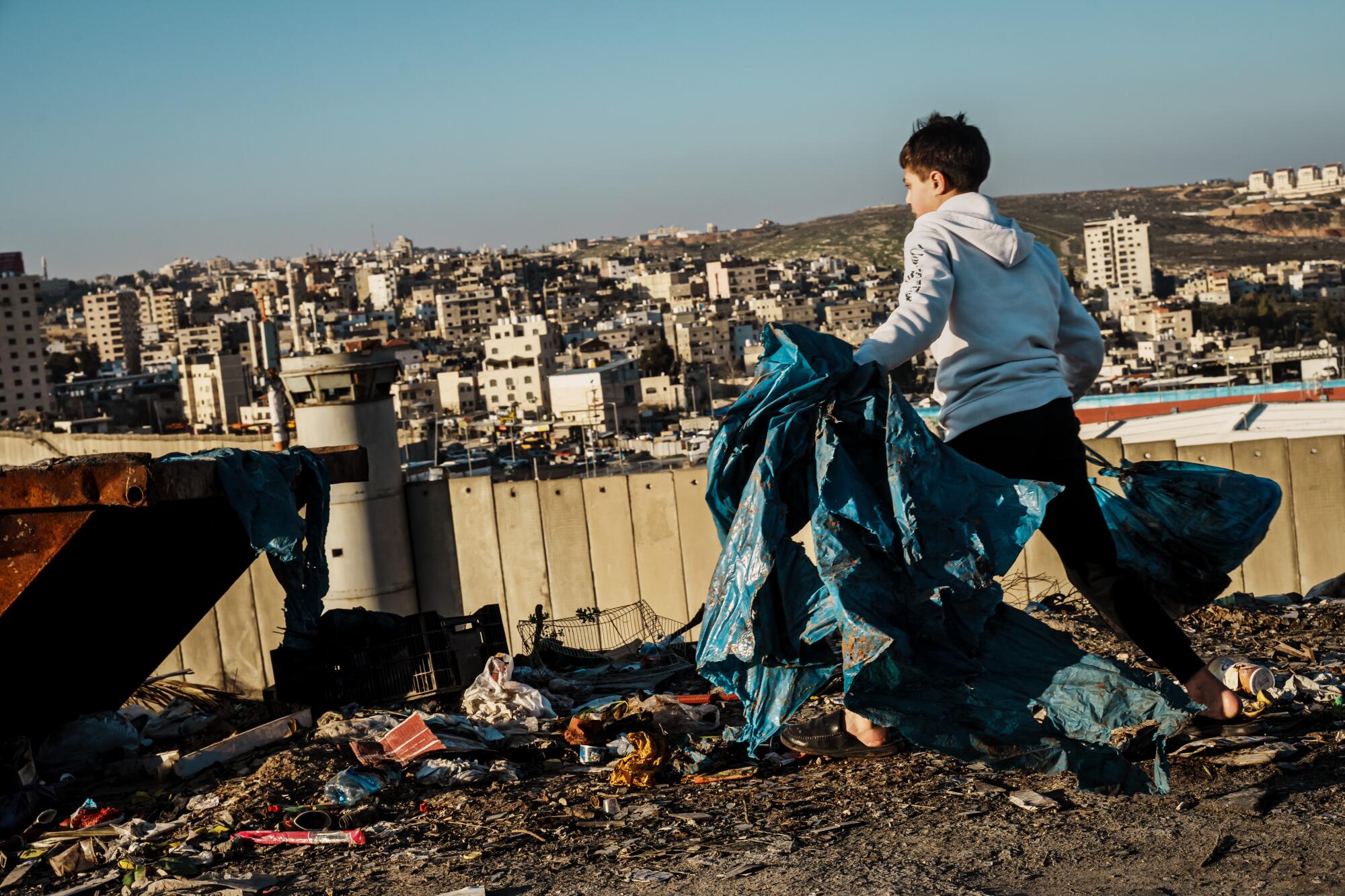 A boy takes out the trash to a bin on a hilltop overlooking the Qalandiya check point.
