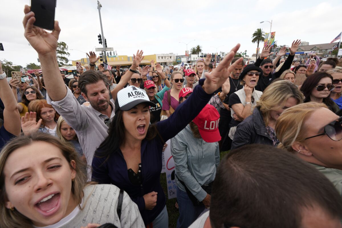 Unmasked people in a crowd shout and lift up their arms.