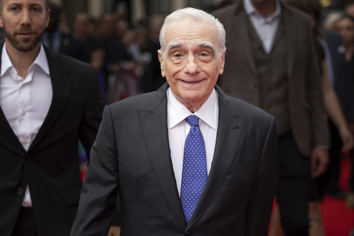 Martin Scorsese, in a dark suit and blue tie, walks the red carpet at a film premiere.