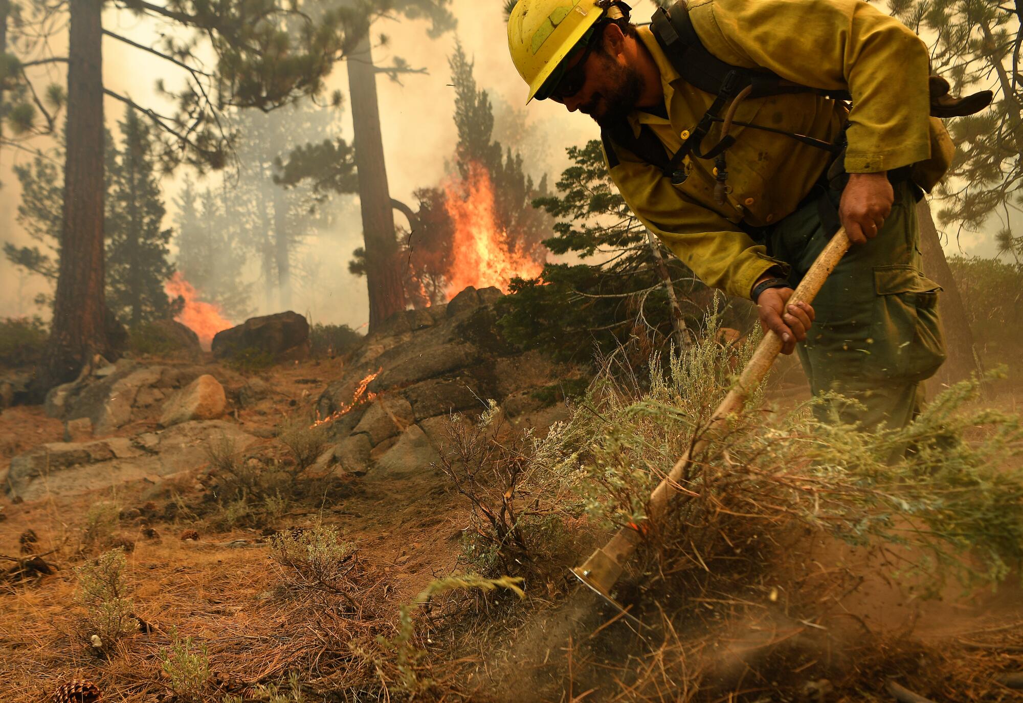 A firefighter uses a hand tool to dig into the dirt as flames burn nearby