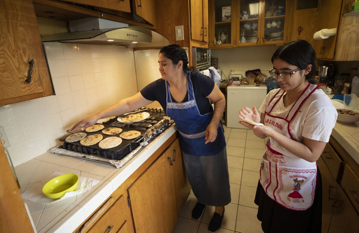 A woman and teenager, in aprons, cook in a kitchen in front of a stovetop grill with rounds of flatbread.