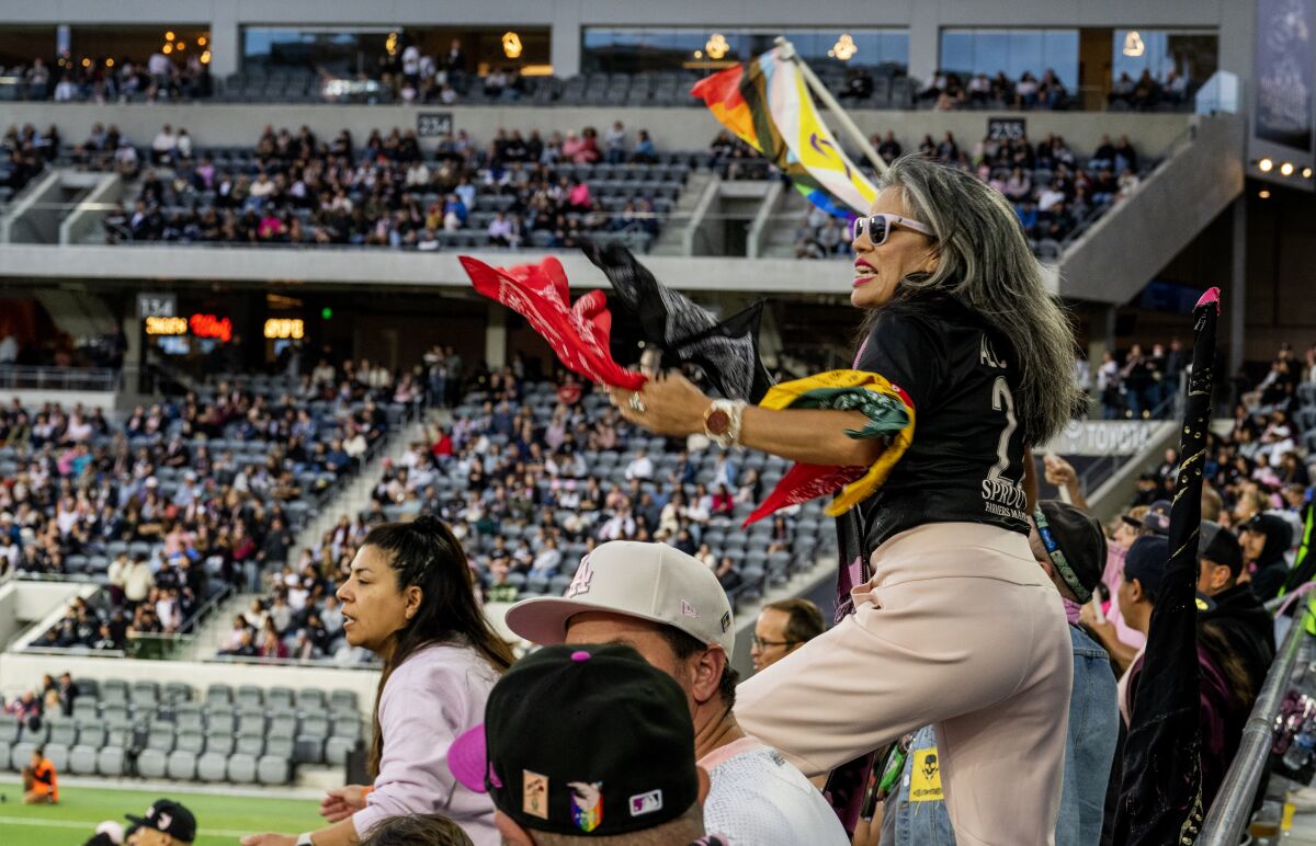 A woman with bandannas tied around her arms stands in a soccer stadium next to others, cheering.