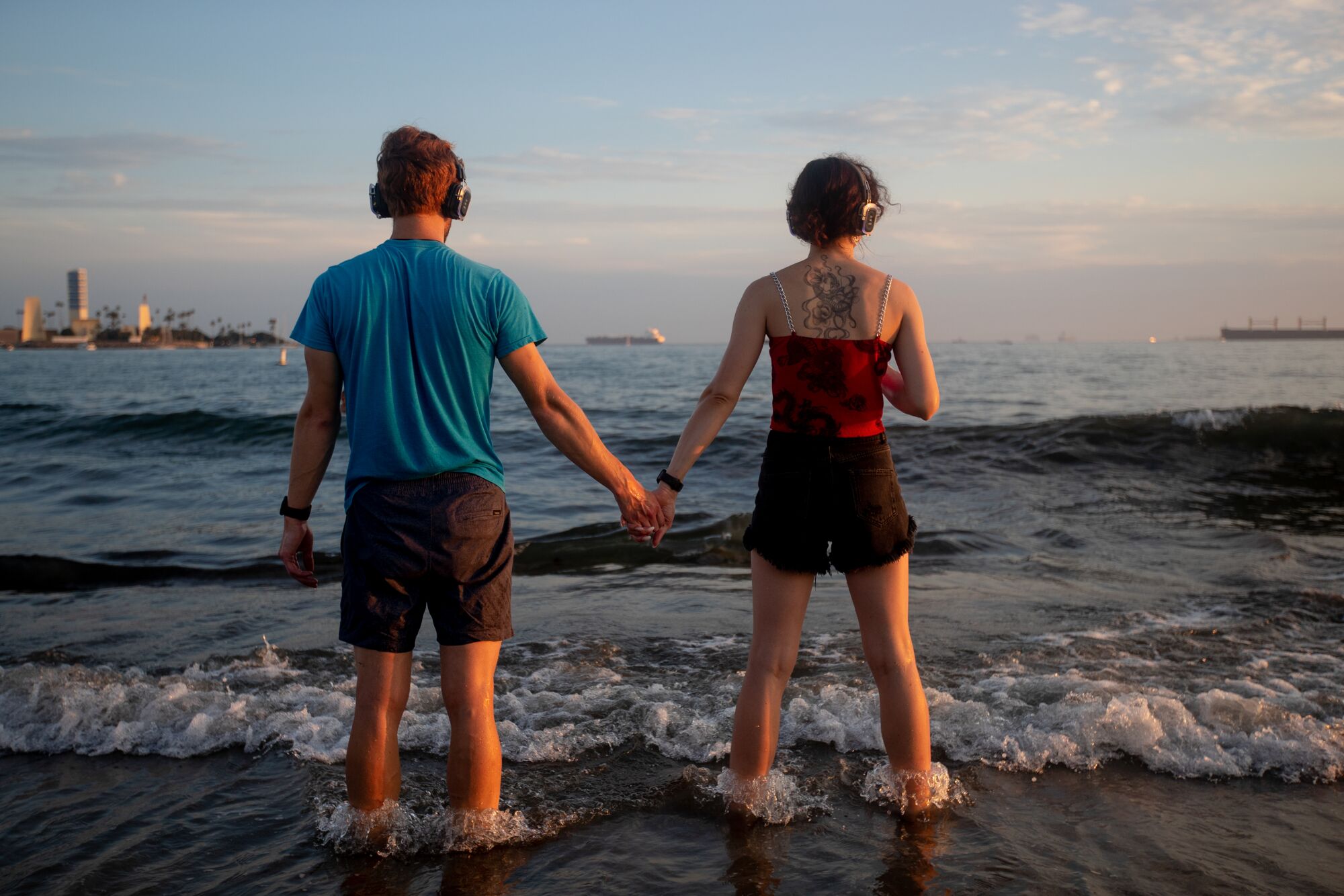 A man and woman standing with their feet in the surf, holding hands and looking out onto the ocean.