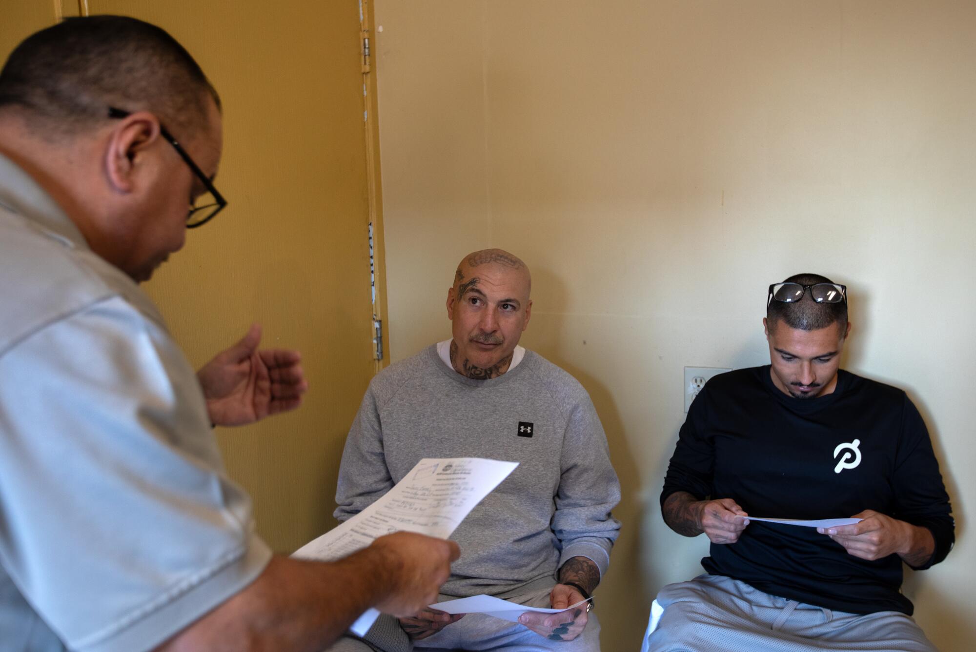 A man reads from a sheet of paper to two others who are seated.