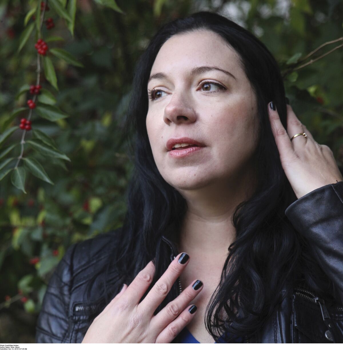 A woman with black hair and black nail polish in front of greenery.
