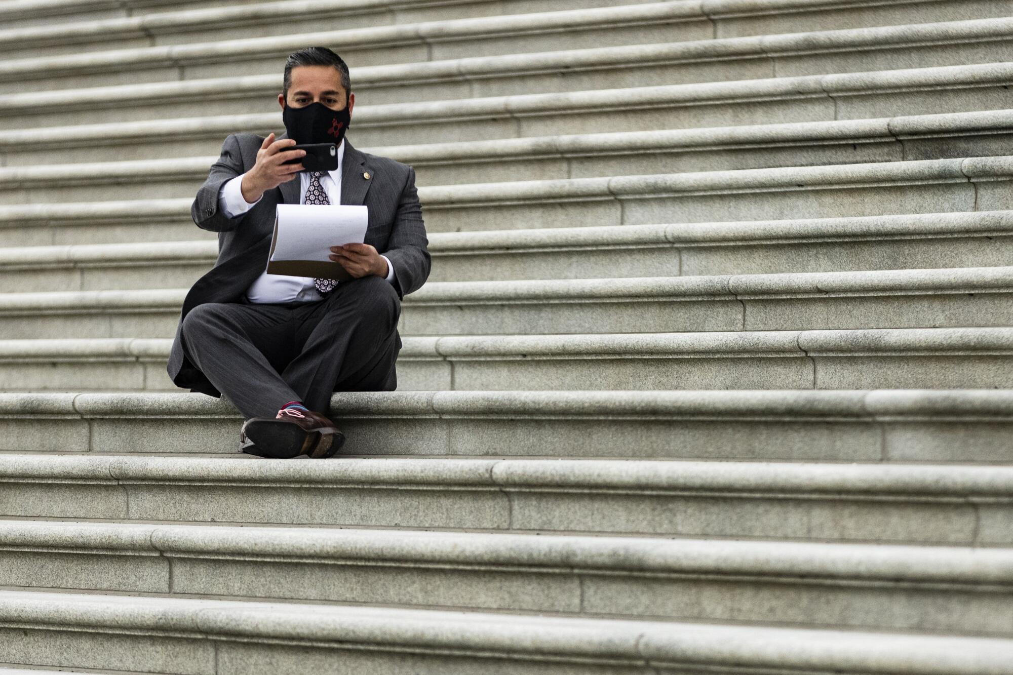 A senator in a mask sits on the Capitol steps while holding up his phone