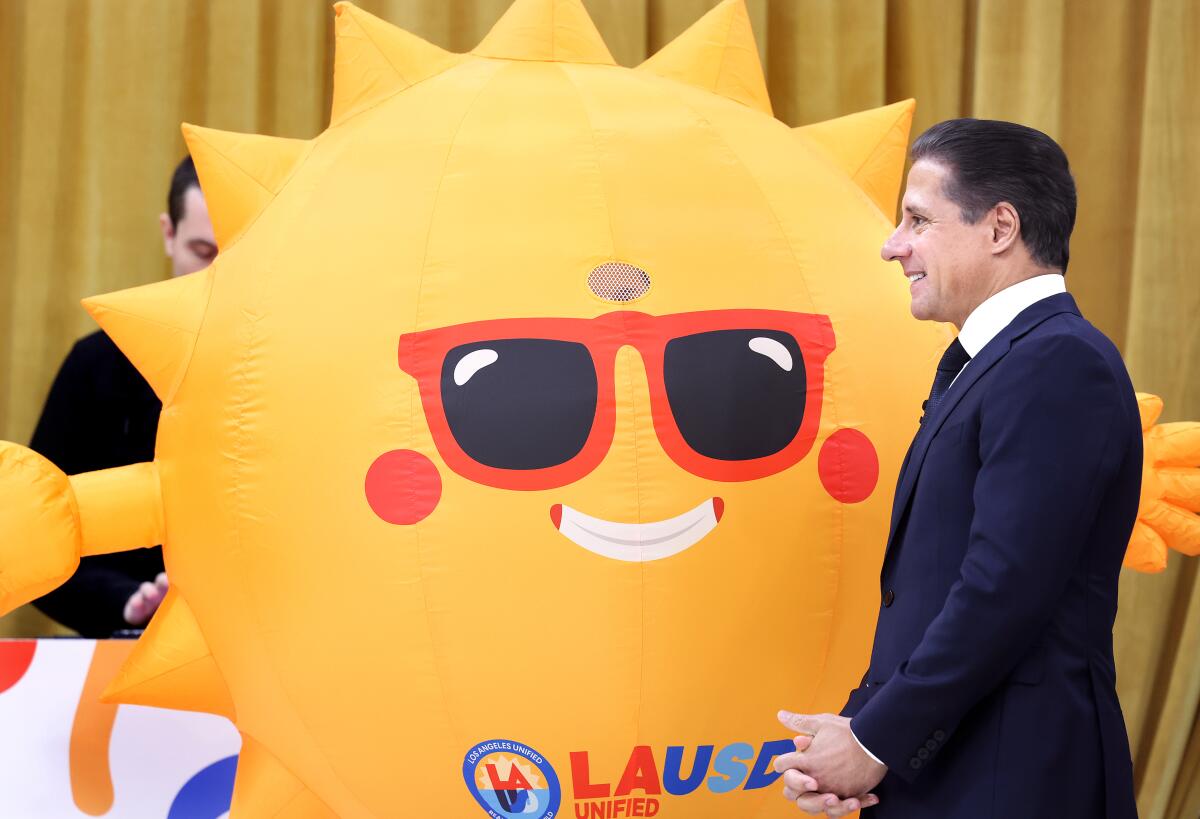 A man with dark hair, in a dark suit and tie, stands with a person wearing a sun costume.