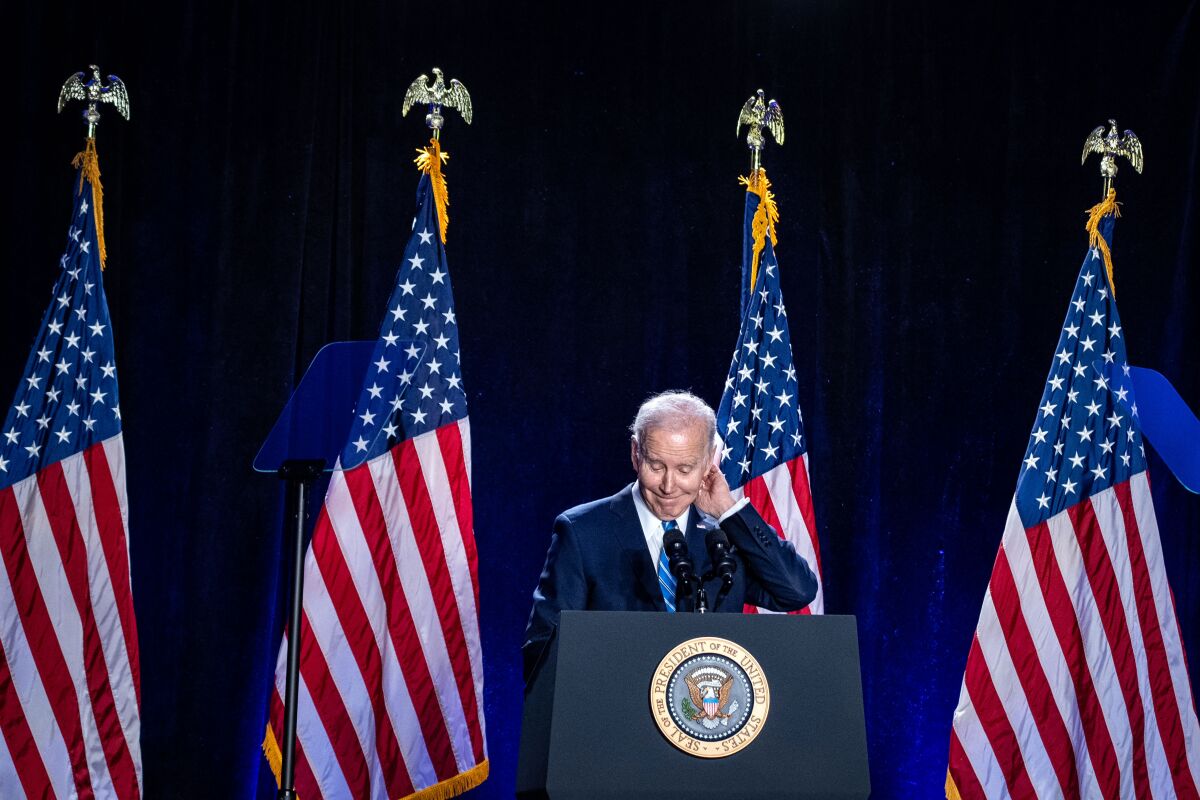 President Biden stands at a lectern with four tall American flags behind him.