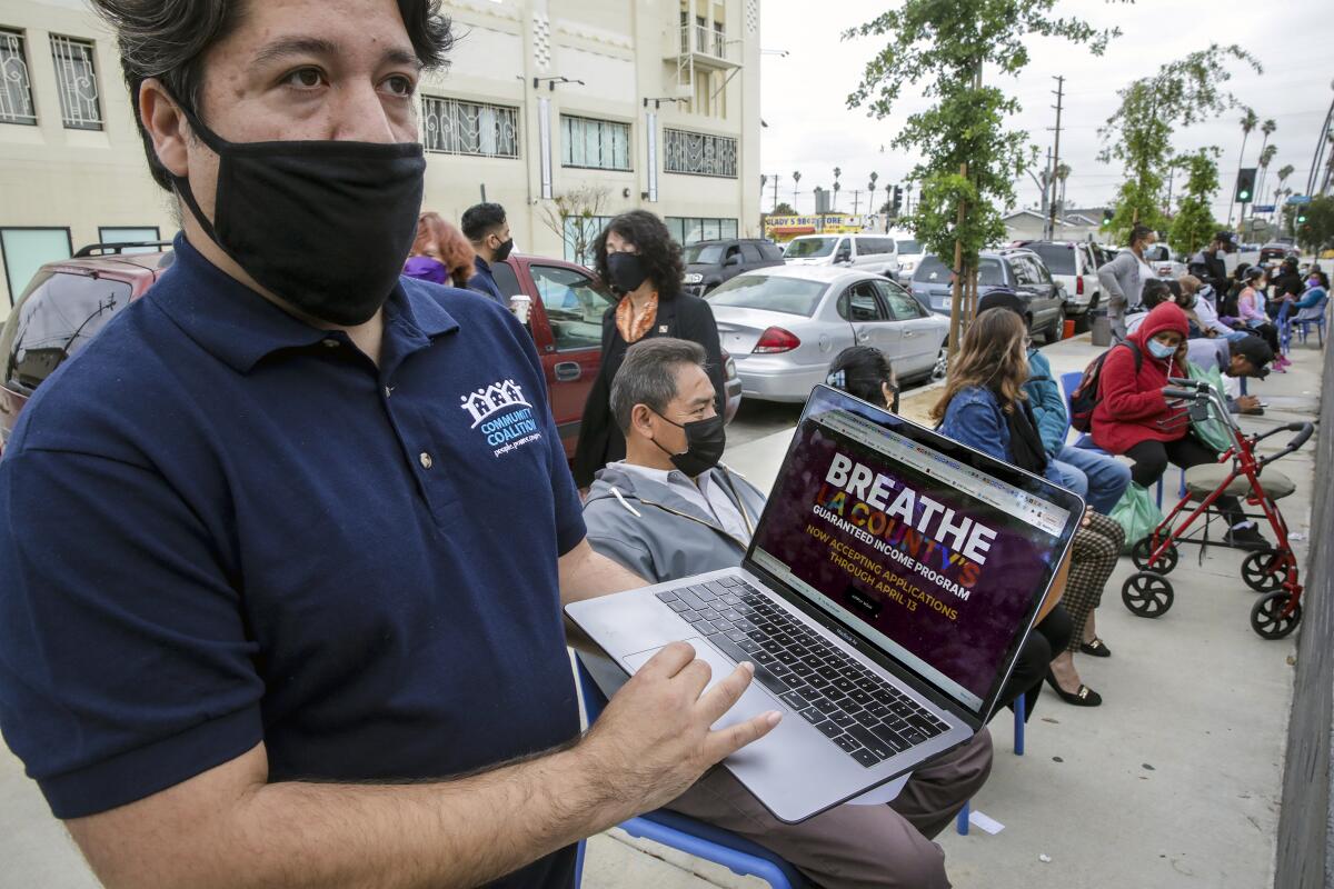A man standing outside near a queue of people holds a laptop displaying the signup page for L.A. County's Breathe program.