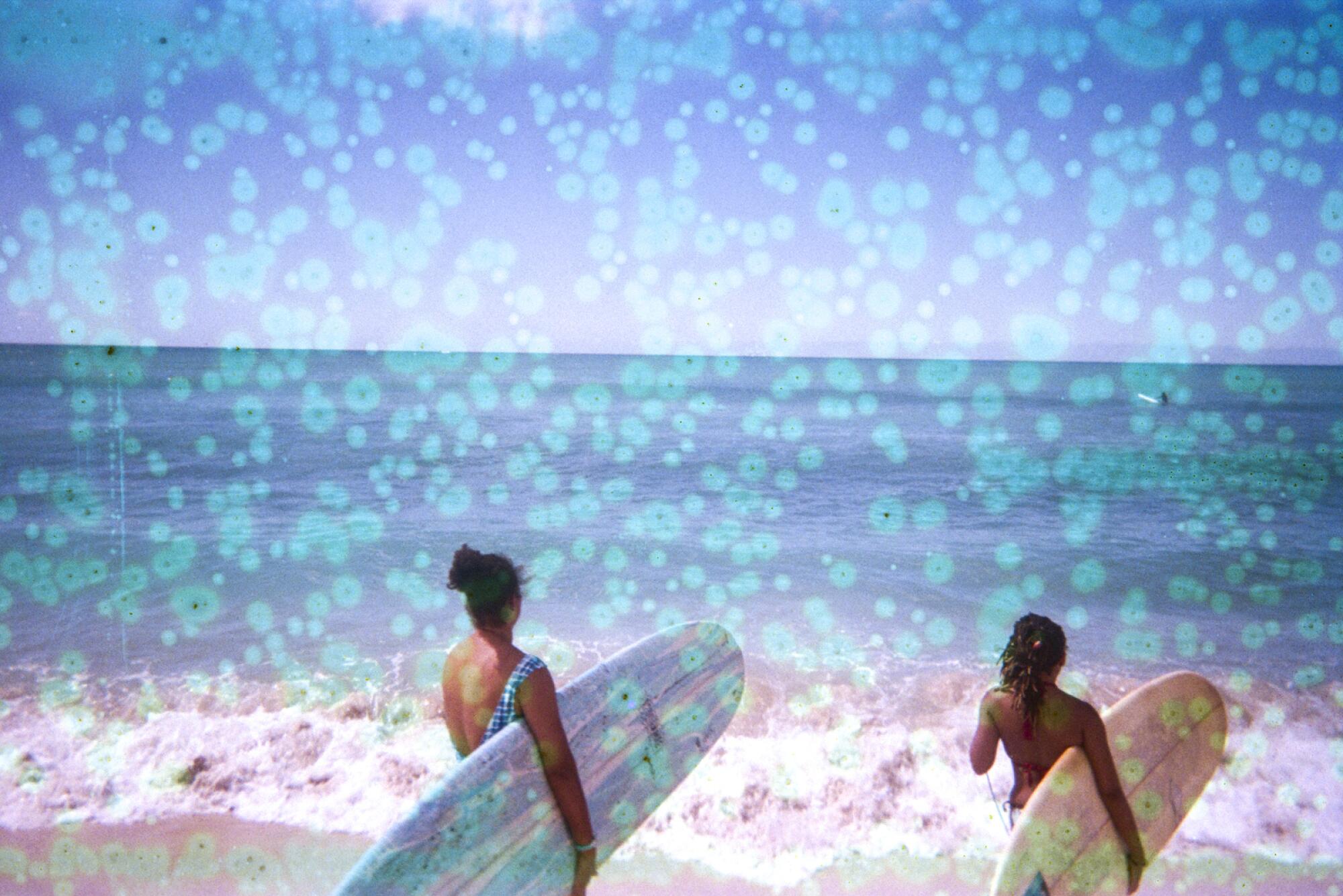 Two surfers carrying their surfboards stand at the edge of the ocean as waves crash onto the beach.