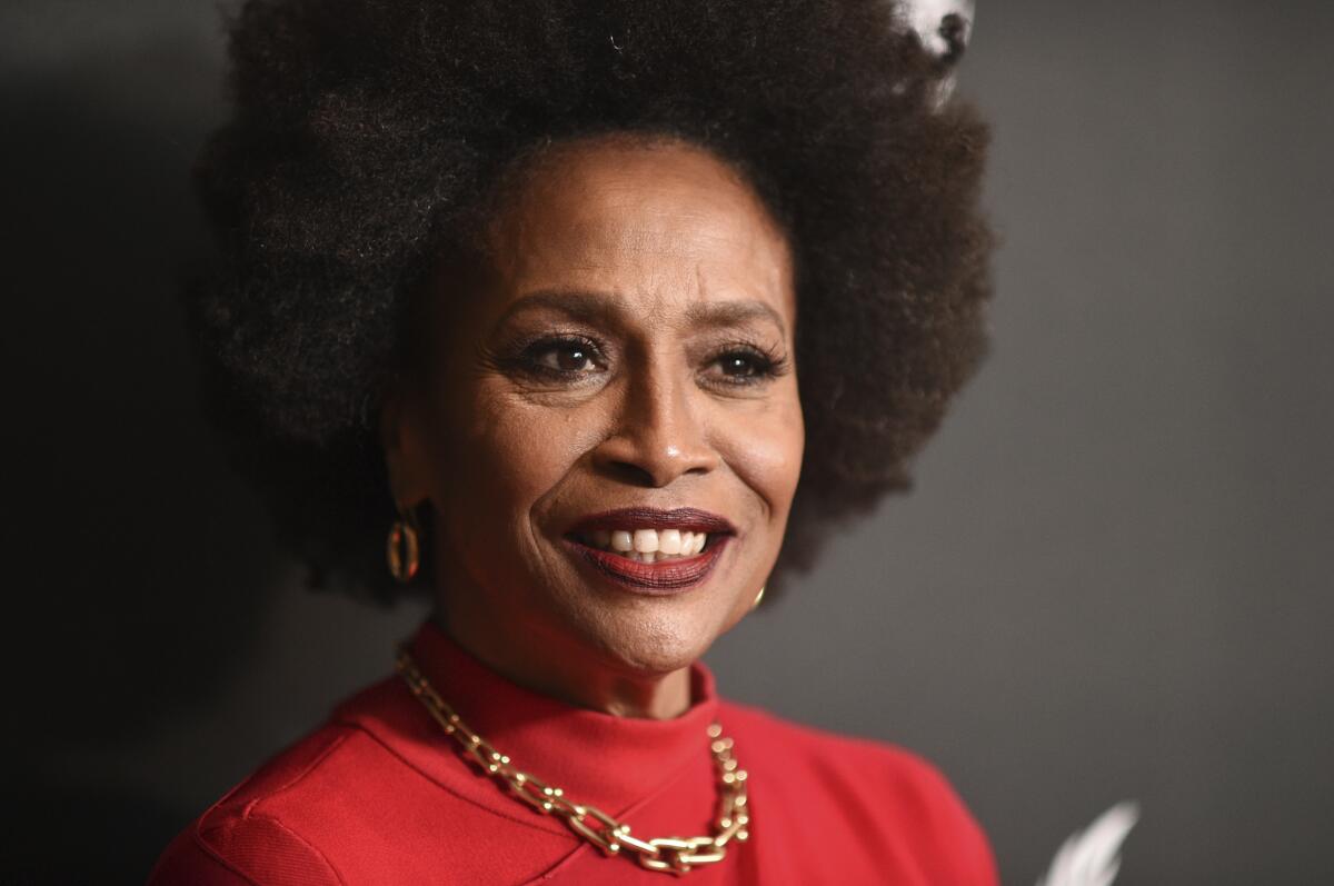 Jenifer Lewis in a red outfit and a gold, thick chain necklace smiling and posing against a black background