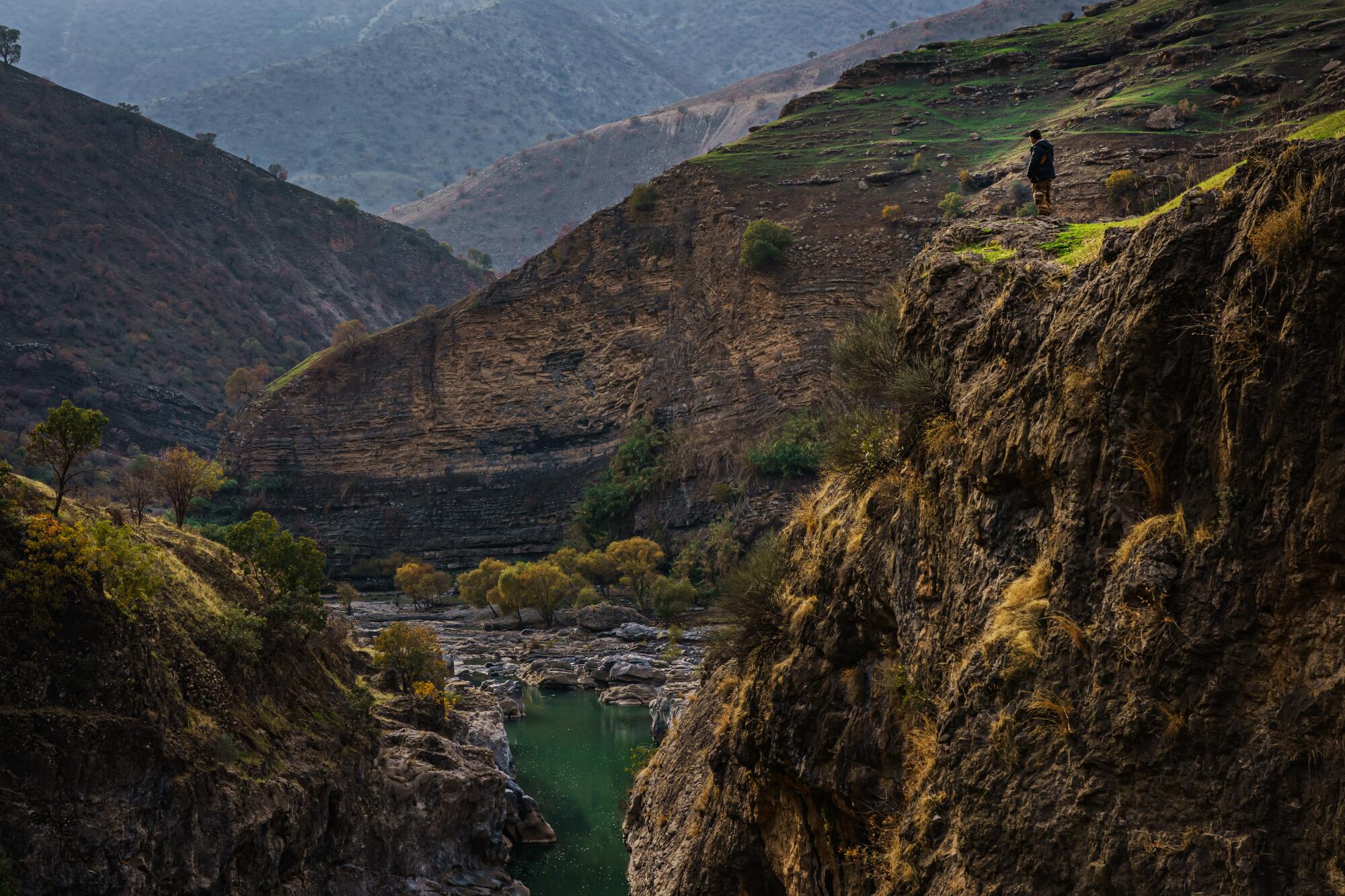 A small figure of a man, right, is seen standing along the edge of a mountain. Far below is a green waterway. 