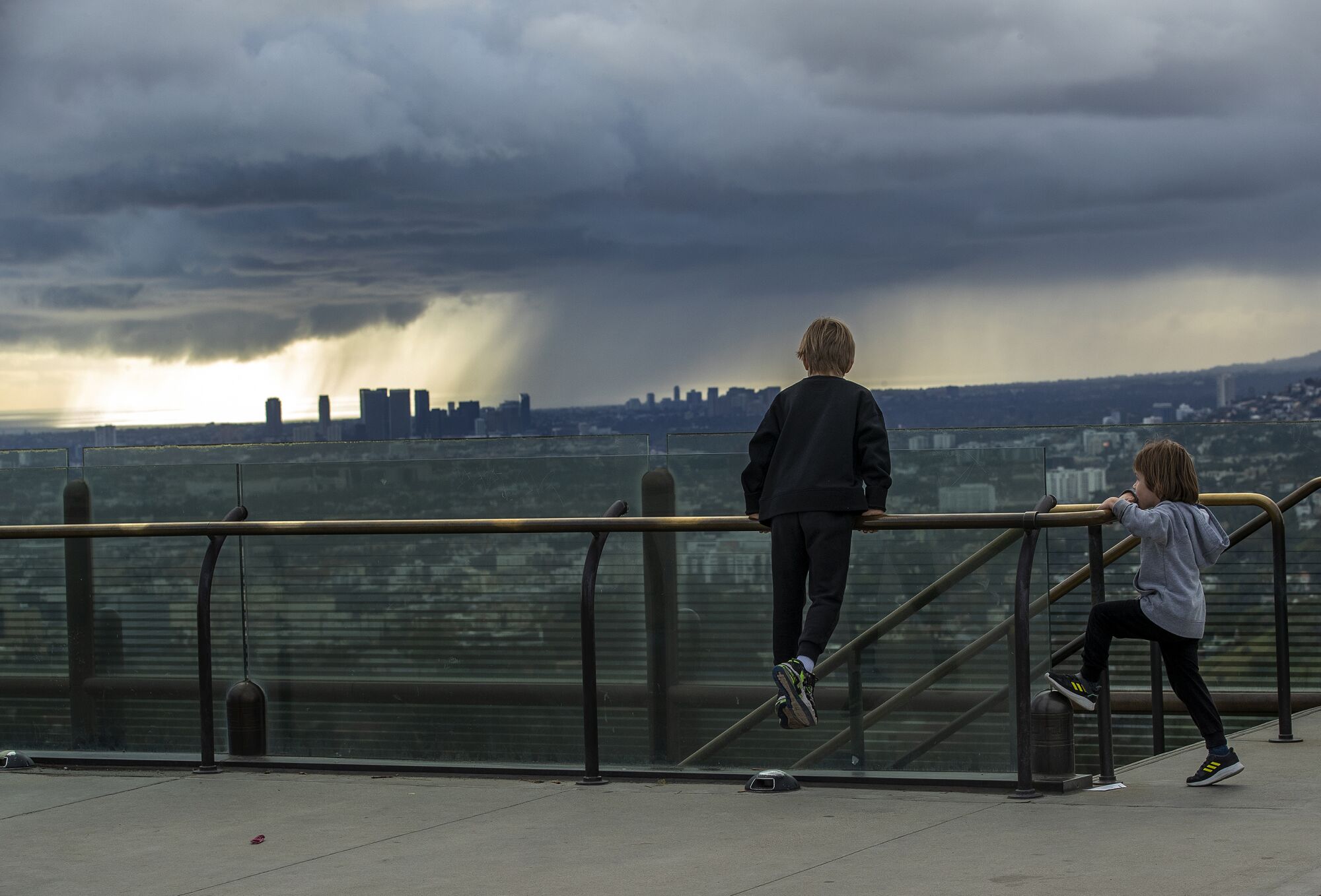 Two young boys climb along railing of a viewing platform. City is in the distance and dark clouds cover the sky.