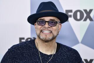 Sinbad in a black wide-brimmed hat, black shirt, necklace with a cross pendant and sunglasses smiling