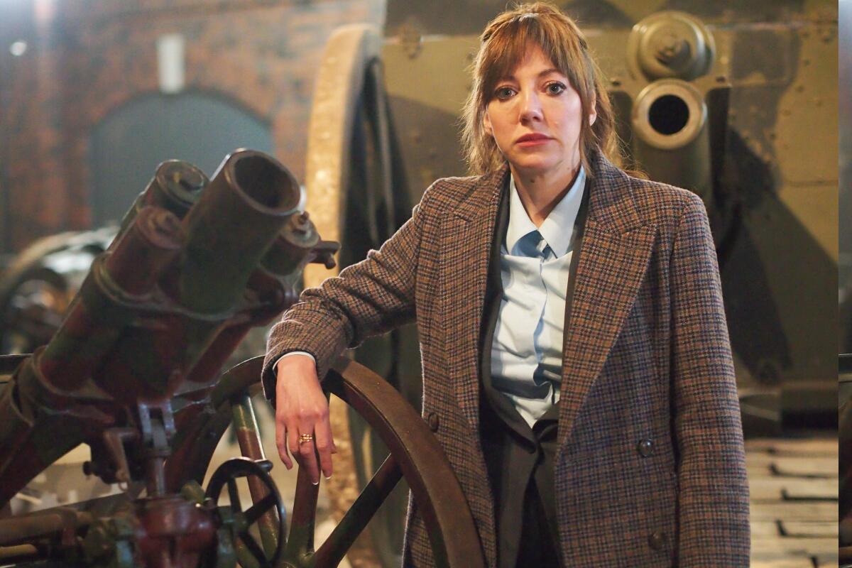 A woman in a tweed jacket leaning on an old cannon looking straight into camera