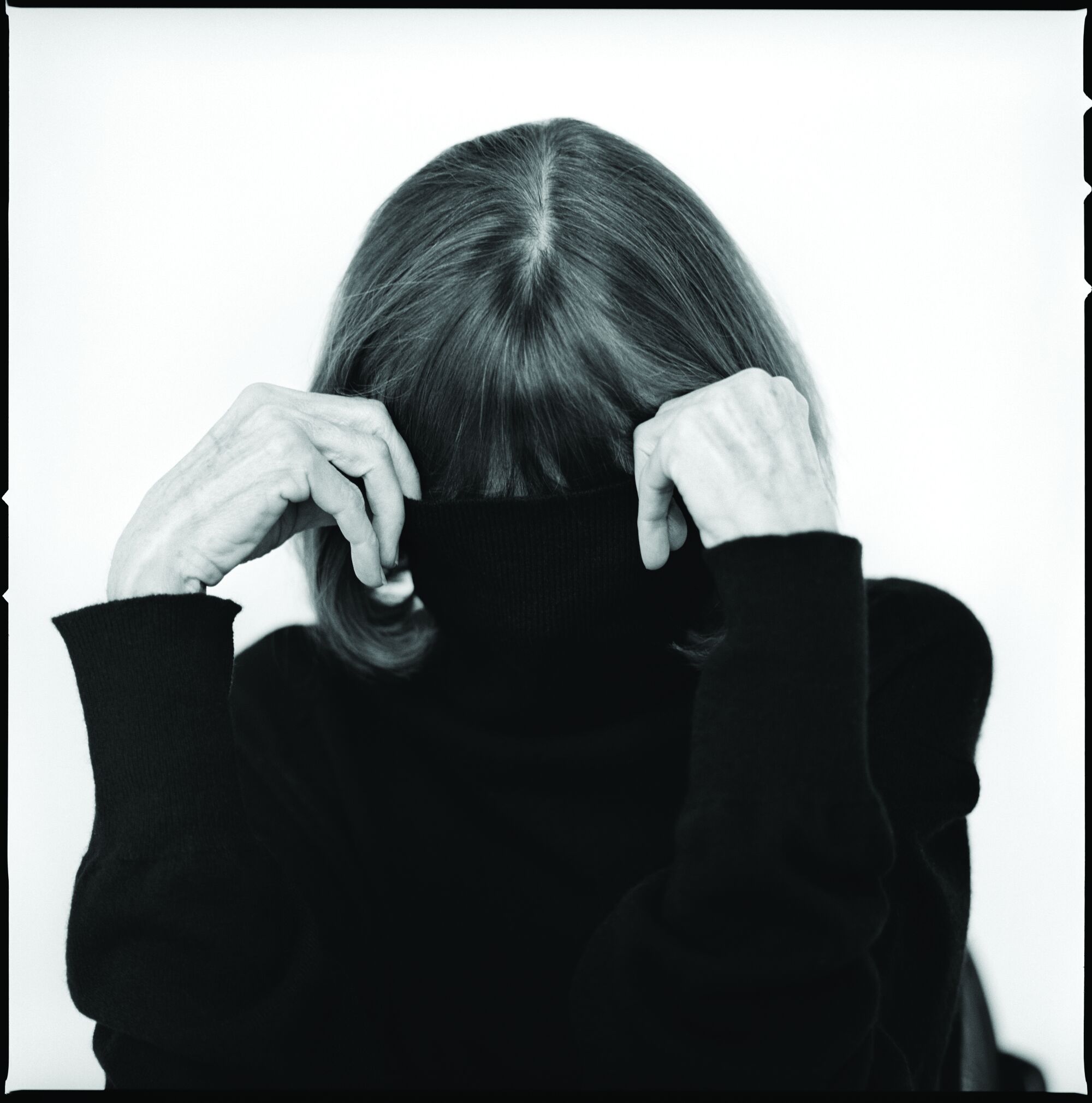 A photograph of Joan Didion pulling a black turtleneck up over her face