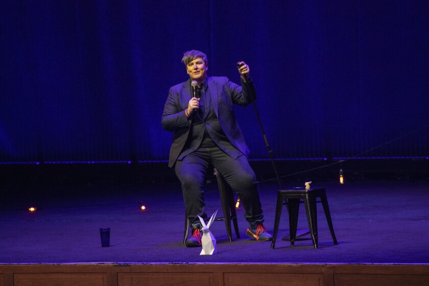 A comedian sitting on a stool onstage with an origami bunny.