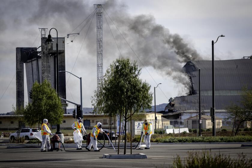 Tustin, CA - November 13: A disaster cleanup crew picks and vacuums up potentially toxic debris from the still-burning WWII-era blimp hangar at the former air base in in Tustin Monday, Nov. 13, 2023. Flare-ups and toxic air from last week's destructive hangar fire in Tustin continue to cause trouble for nearby residents. The City of Tustin took to X, formerly Twitter, to confirm that the western wall of the 17-story building reignited Sunday night. Orange County Fire Authority personnel remained on the scene keeping watch of the blaze on Monday morning, with one firefighter telling KTLA 5's Annie Rose Ramos that all they could do was let it burn out. (Allen J. Schaben / Los Angeles Times)