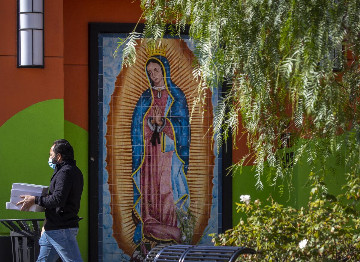 A man carries boxes of baked goods and passes by a mural of Our Lady of Guadalupe in downtown Santa Ana on Wednesday.