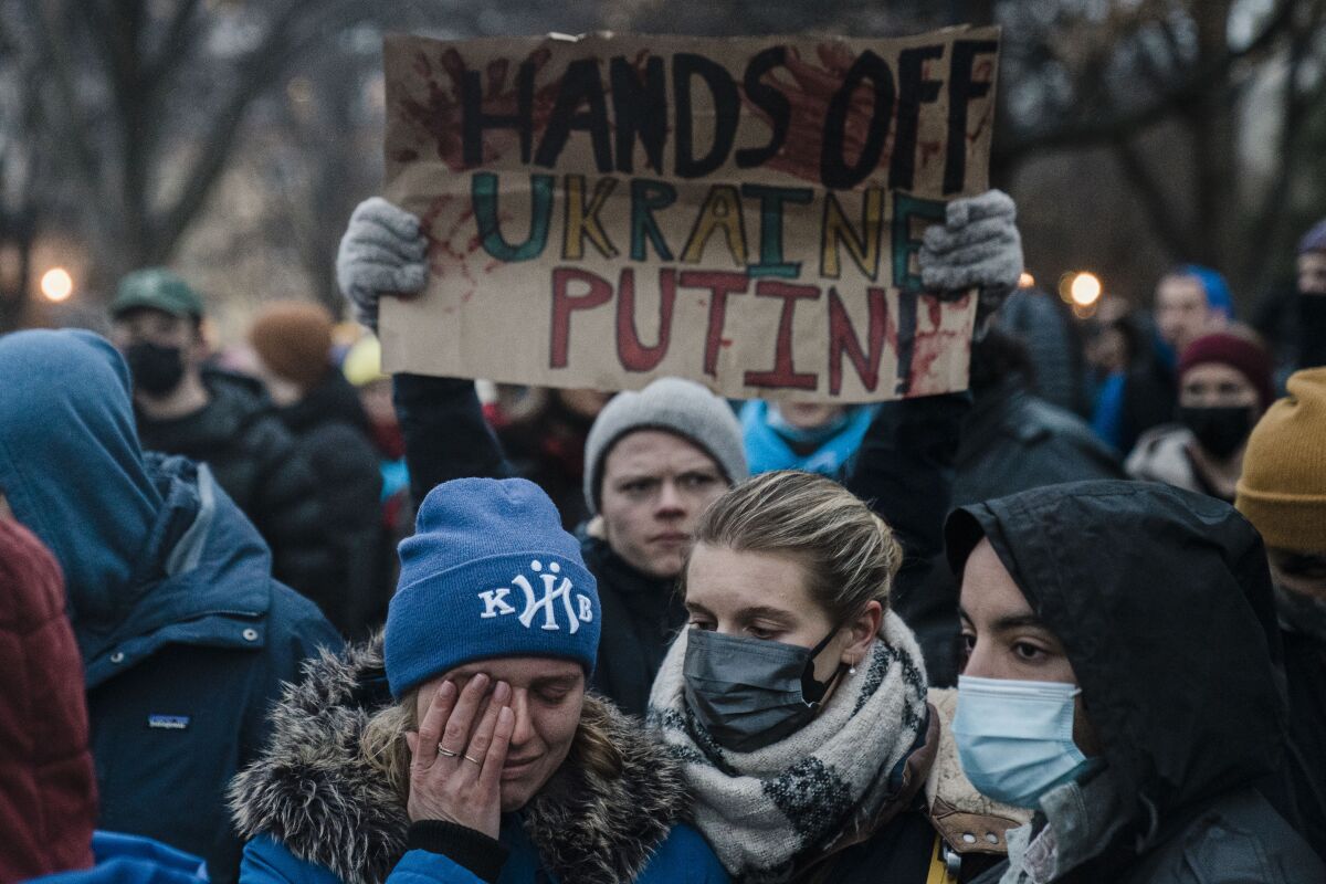 A protester in a crowd holds up a sign saying "Hands off Ukraine Putin," while a woman brushes tears away