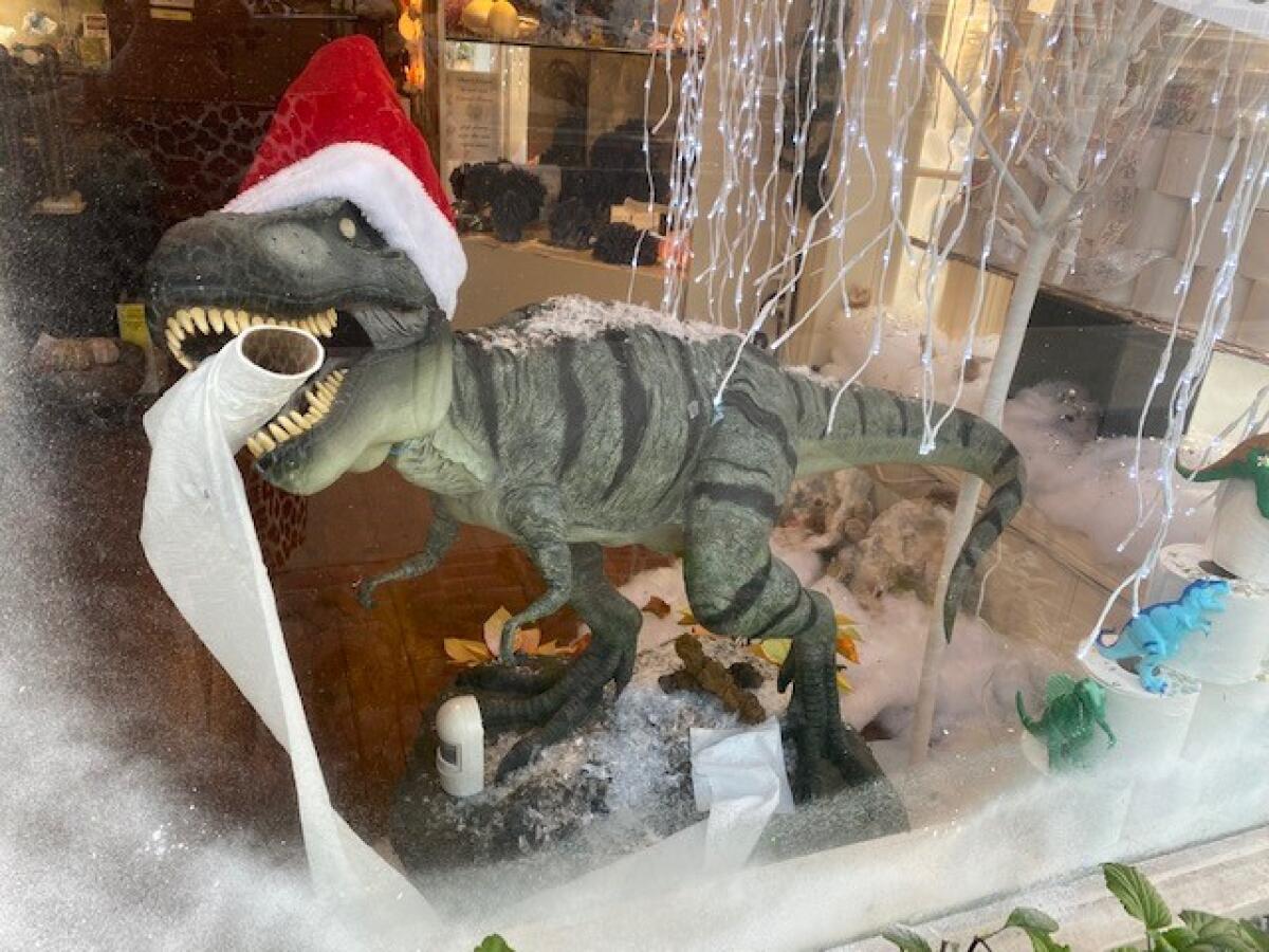 A T-Rex dinosaur holds toilet paper in its jaw in KRISTALLE's holiday window decoration display.