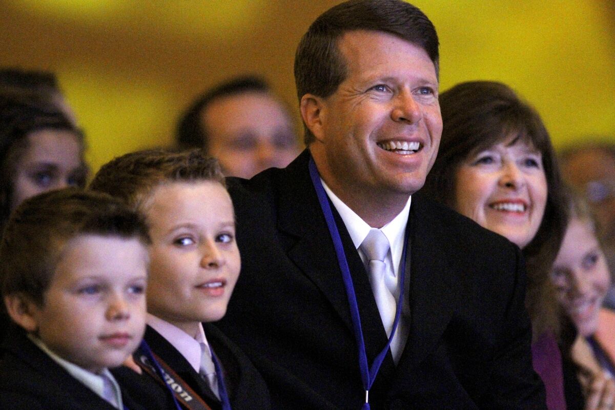 Jim Bob Duggar sits next to two of his sons, all in suits, and looks toward a stage.