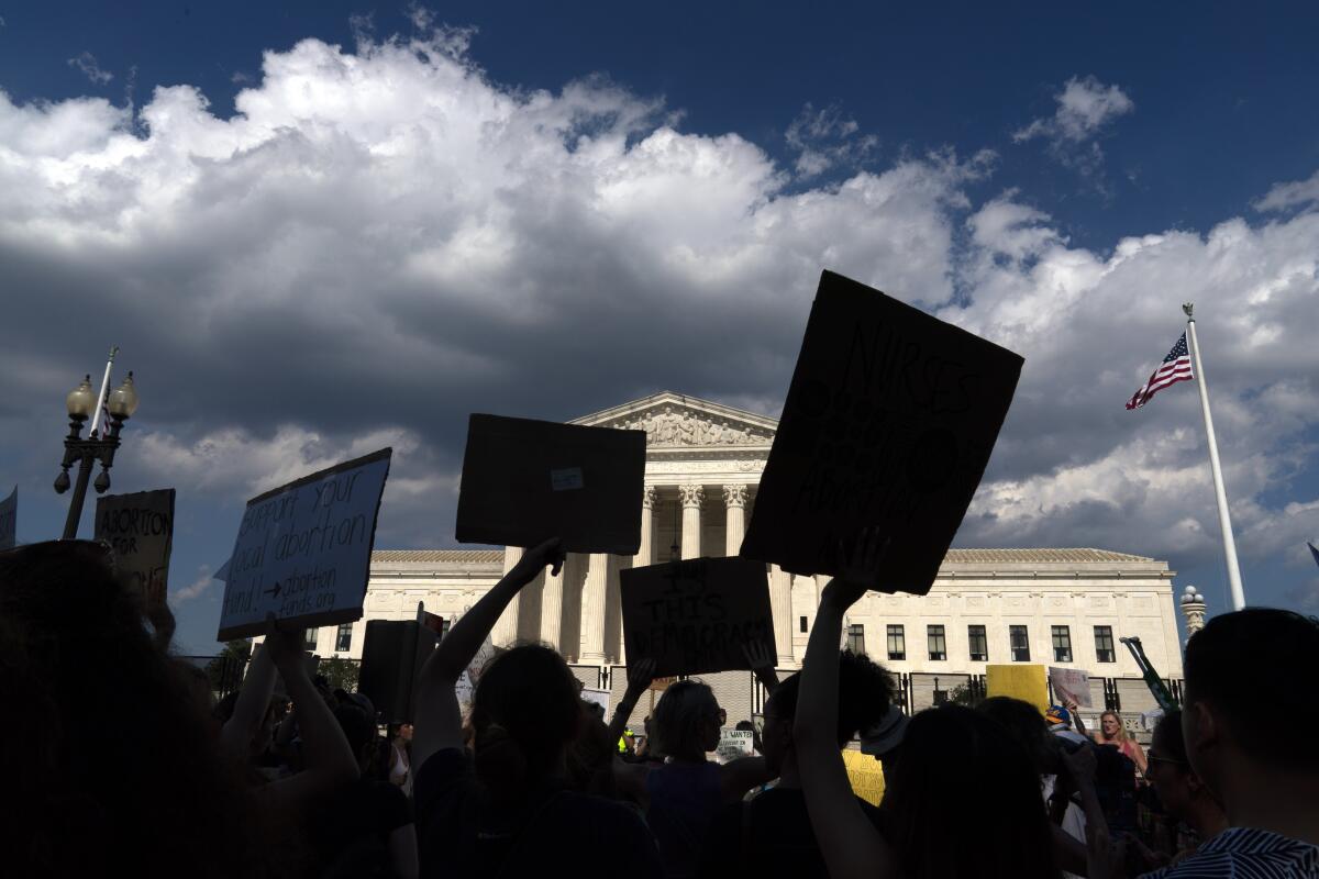 Demonstrators raising signs, seen in silhouette against the U.S. Supreme Court building and a partially cloudy sky 