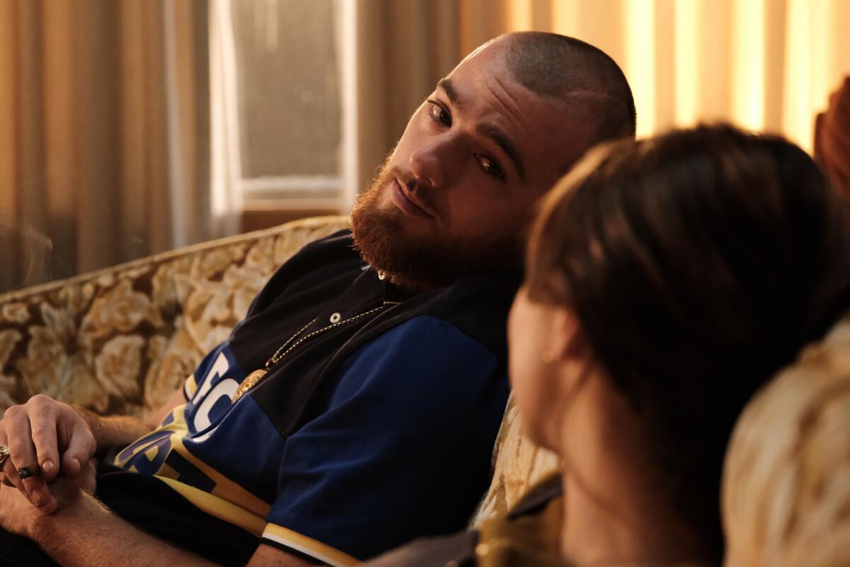 A young man with a shaved head and beard leans back on a couch, looking at his girlfriend.