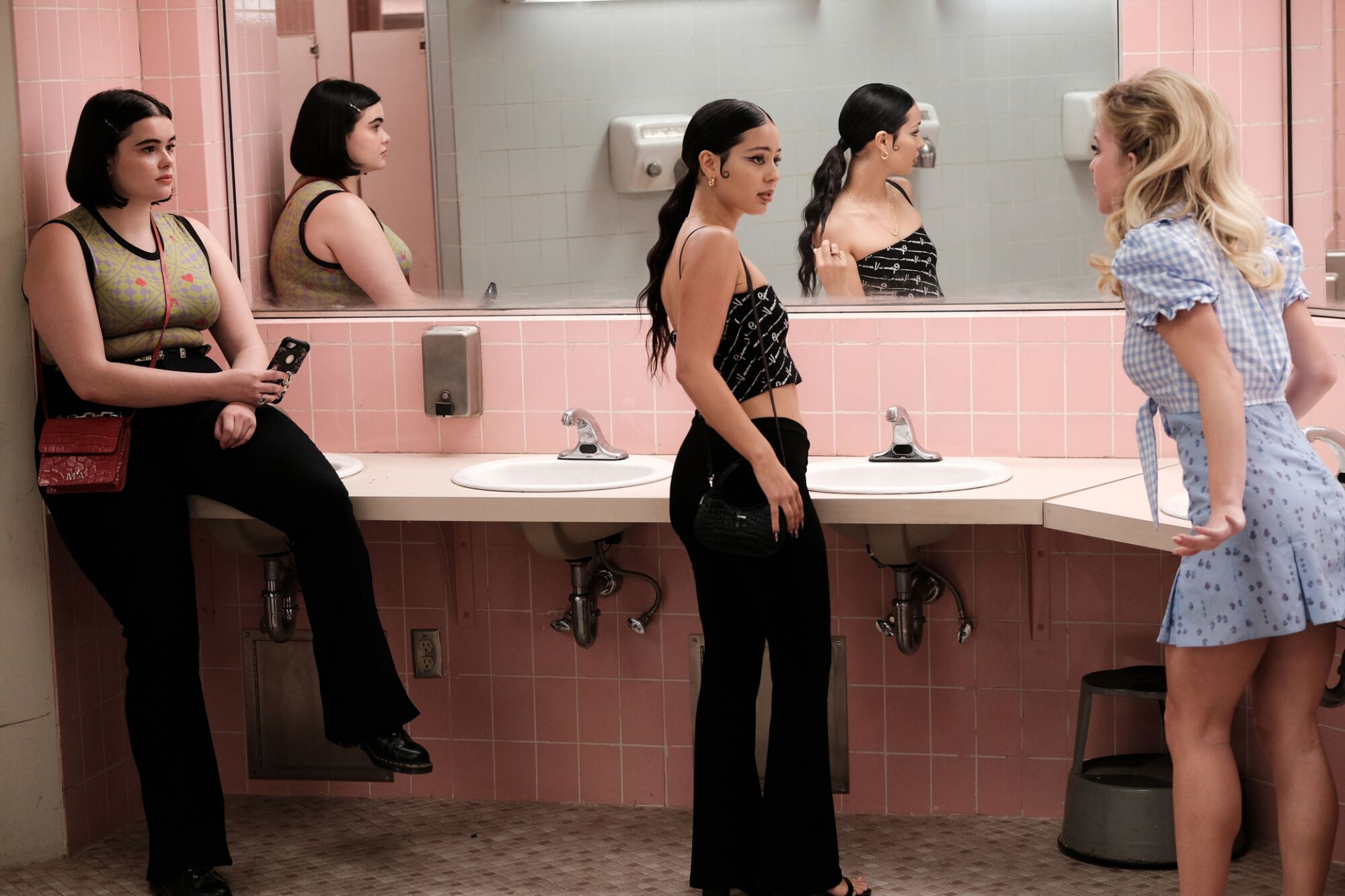 Three women and their reflections in the over-sink mirror of a public bathroom with pink tile.