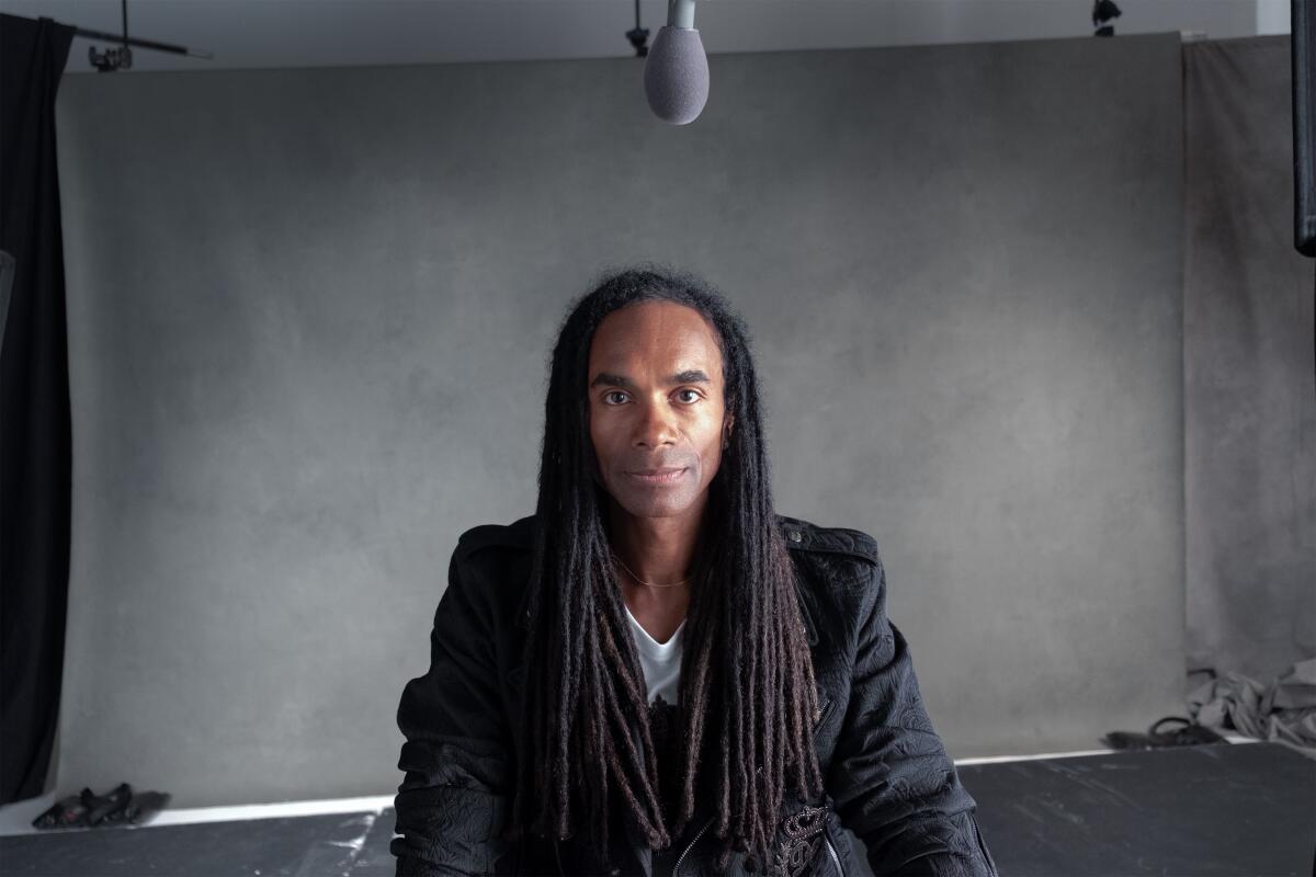 Fab Morvan sits in front of gray backdrop in a white shirt and black jacket with his dreadlocks over his chest.