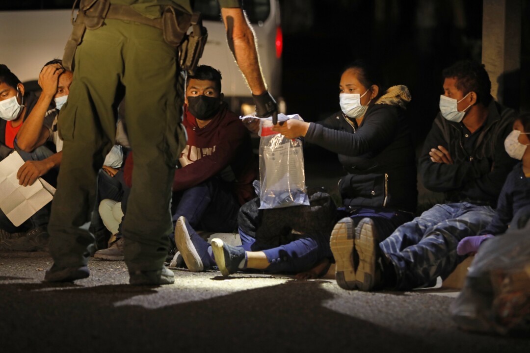 A Border Patrol agent bends over to take a plastic bag of personal items from a migrant woman sitting on the ground
