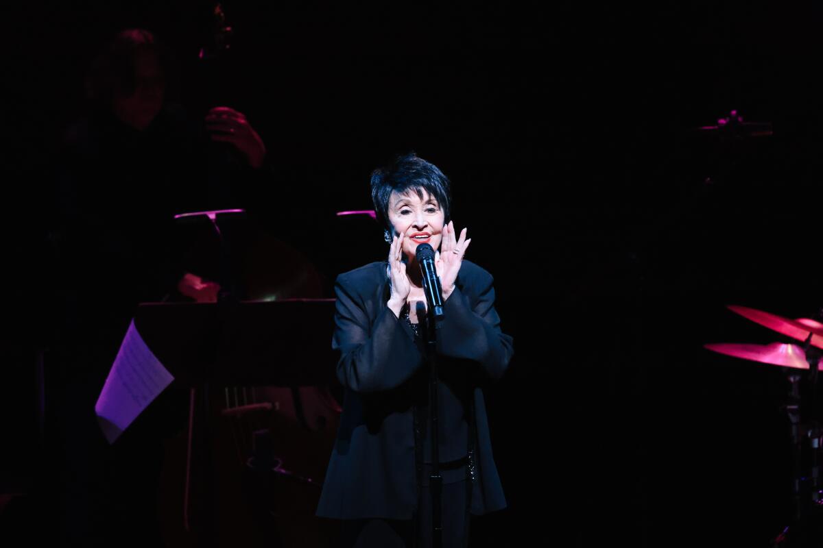 Chita Rivera, dressed in black, sings expressively into a microphone on a darkened stage.