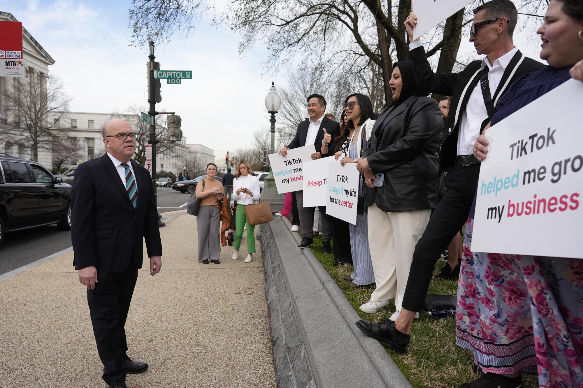 人们在美国国会大厦附近的人行道上排起队伍，举着支持 TikTok 的标语，旁边是一名西装革履的男子