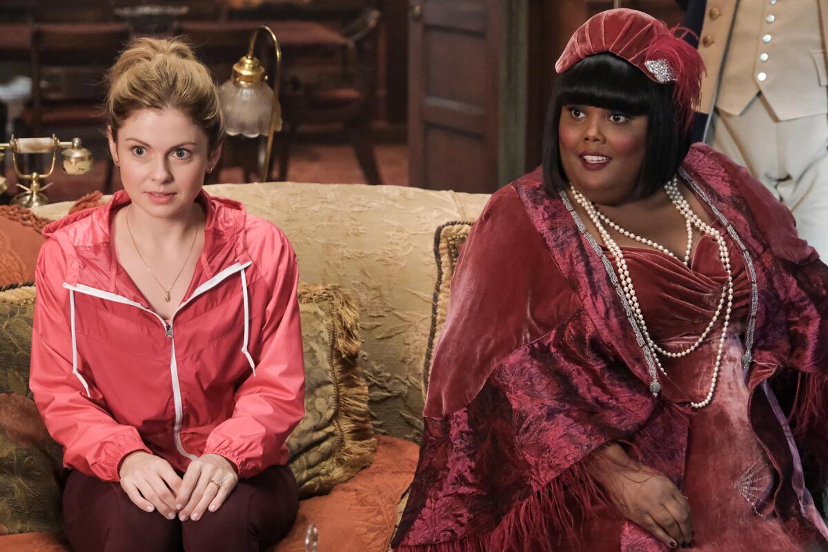 A woman in a tracksuit sits next to a woman in a velvet dress, shawl and hat.