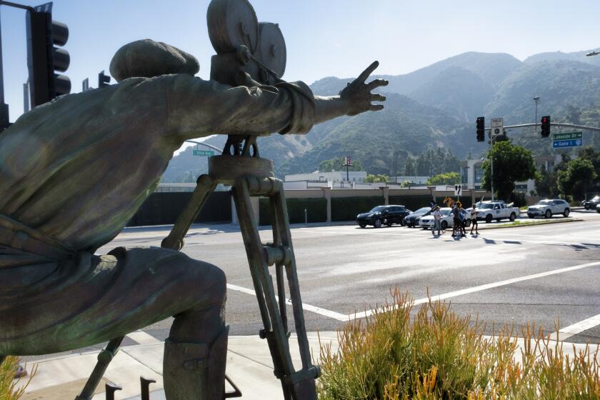 With a statue of a cameraman in the foreground, SAG-AFTRA picketers carrying signs cross a street near the gates of Warner Bros. studios in Burbank, Calif., Tuesday, Sep. 26, 2023. (AP Photo/Richard Vogel)