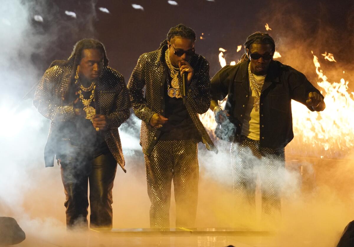 Three men in black outfits performing on a smoky stage with flames in the background