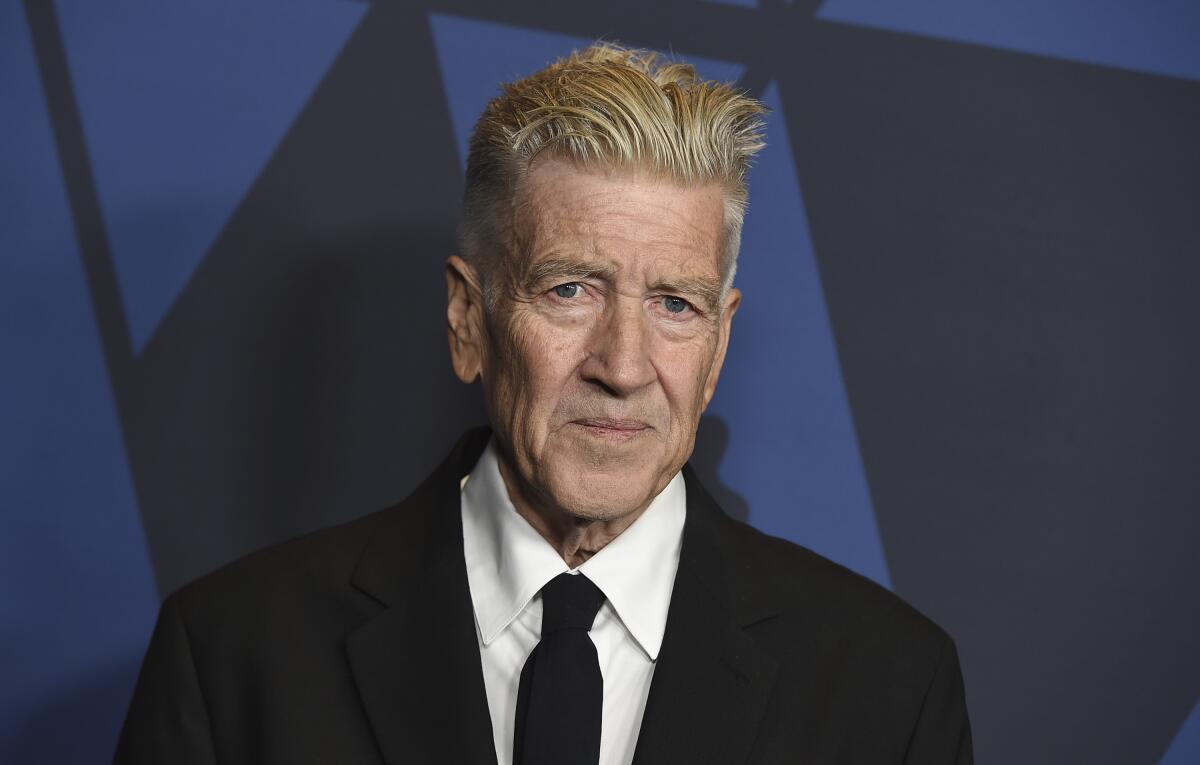 David Lynch looking tight-lipped while posing in a black jacket and tie and white collared shirt 