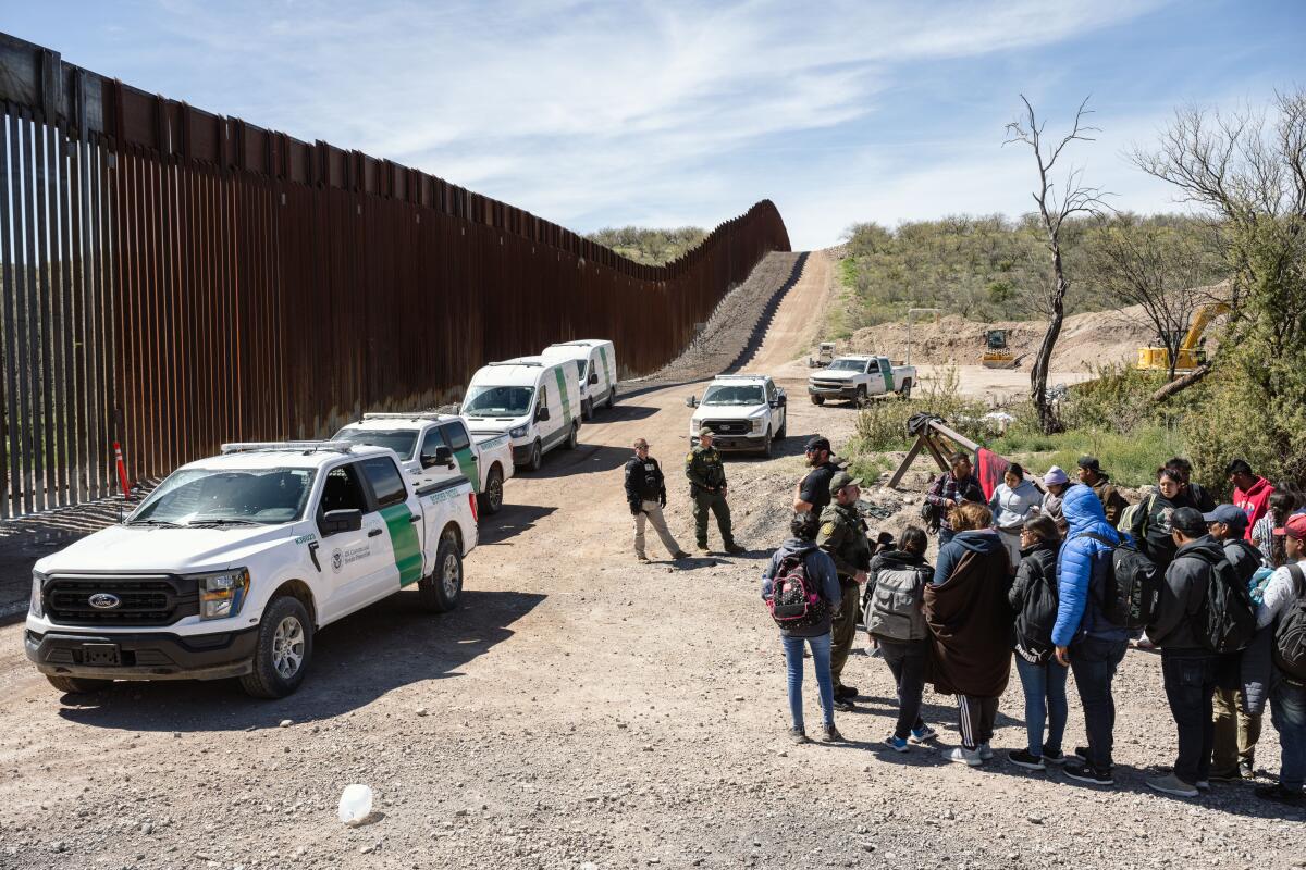 Several white trucks parked along a border wall as agents in uniform talk with over a dozen people, many with backpacks