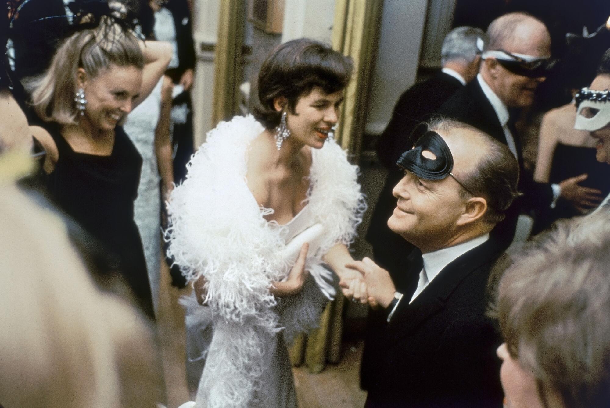 Truman Capote, with a mask resting on his forehead, greets elegantly dressed party guests.