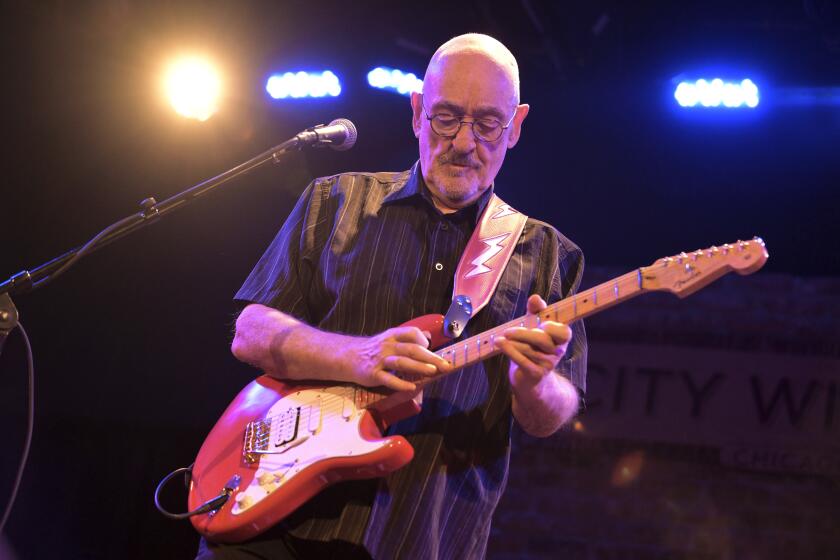 Dave Mason in a black button-down shirt playing a guitar on a stage 