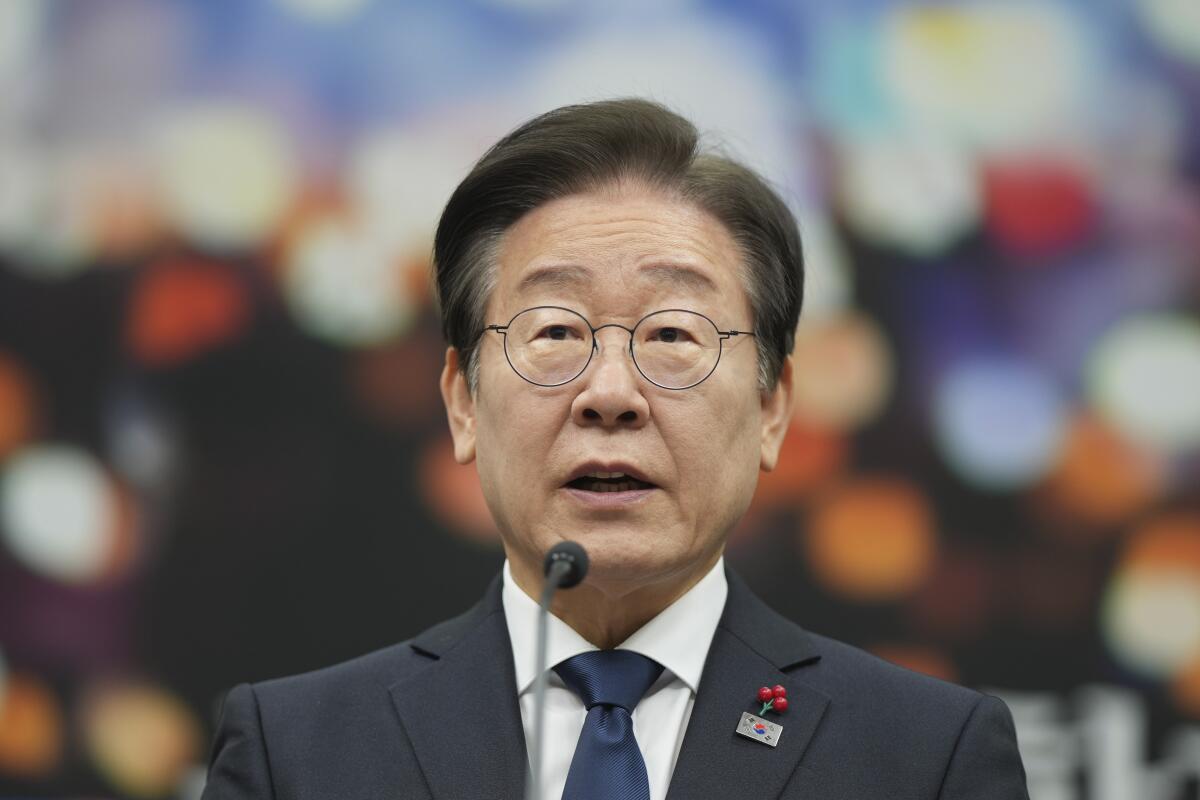 A man with dark hair, in glasses, dark suit and tie, speaks before a microphone