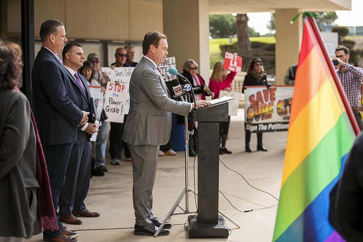 A man in a suit stands outdoors at a lectern, speaking into a microphone with people lined up behind him.