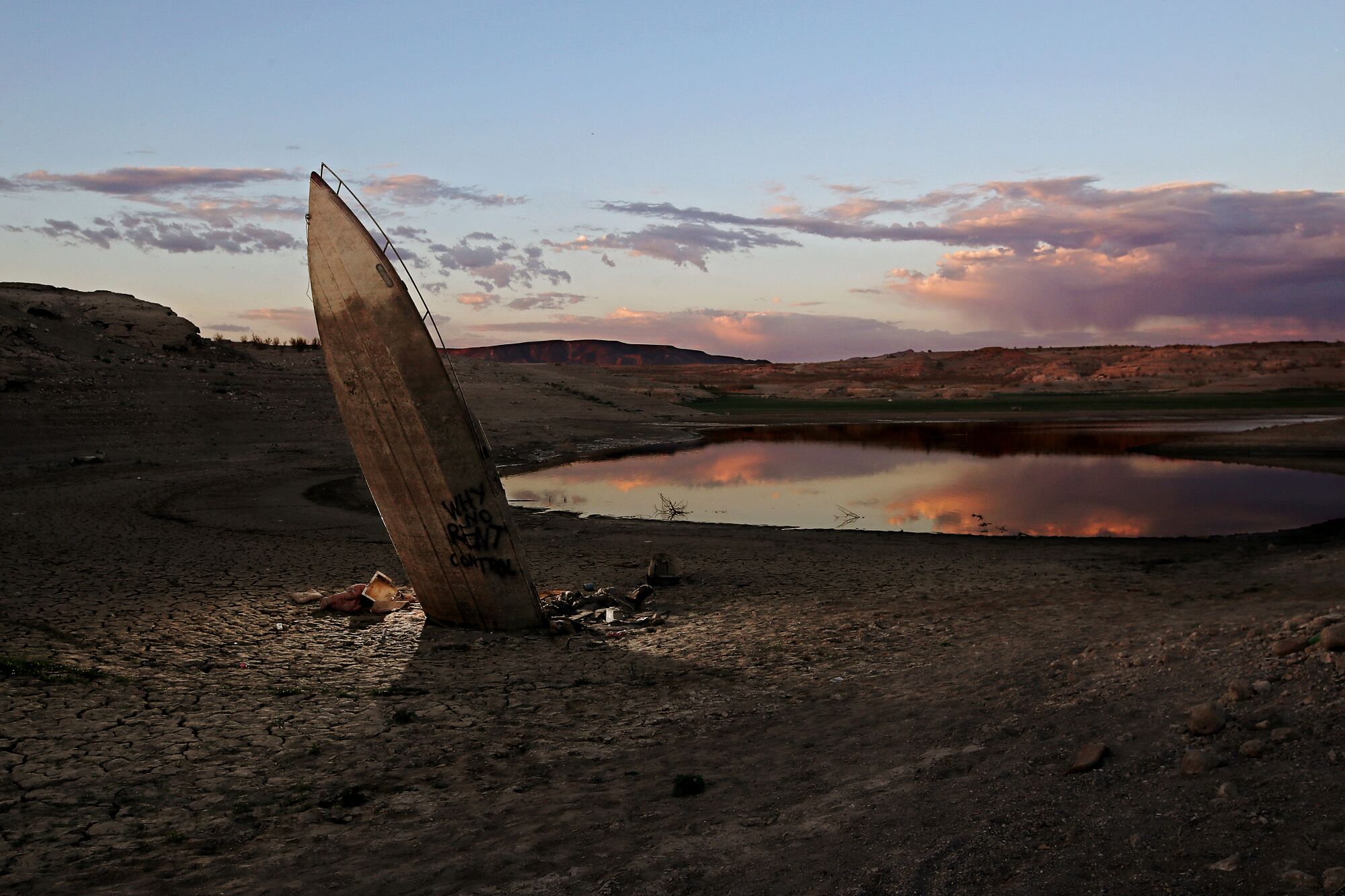 The decaying hull of a speedboat stands perpendicular from exposed lake bed. 