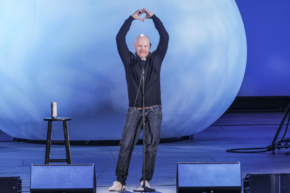 Bald man in shirt and jeans on stage with hands in the air