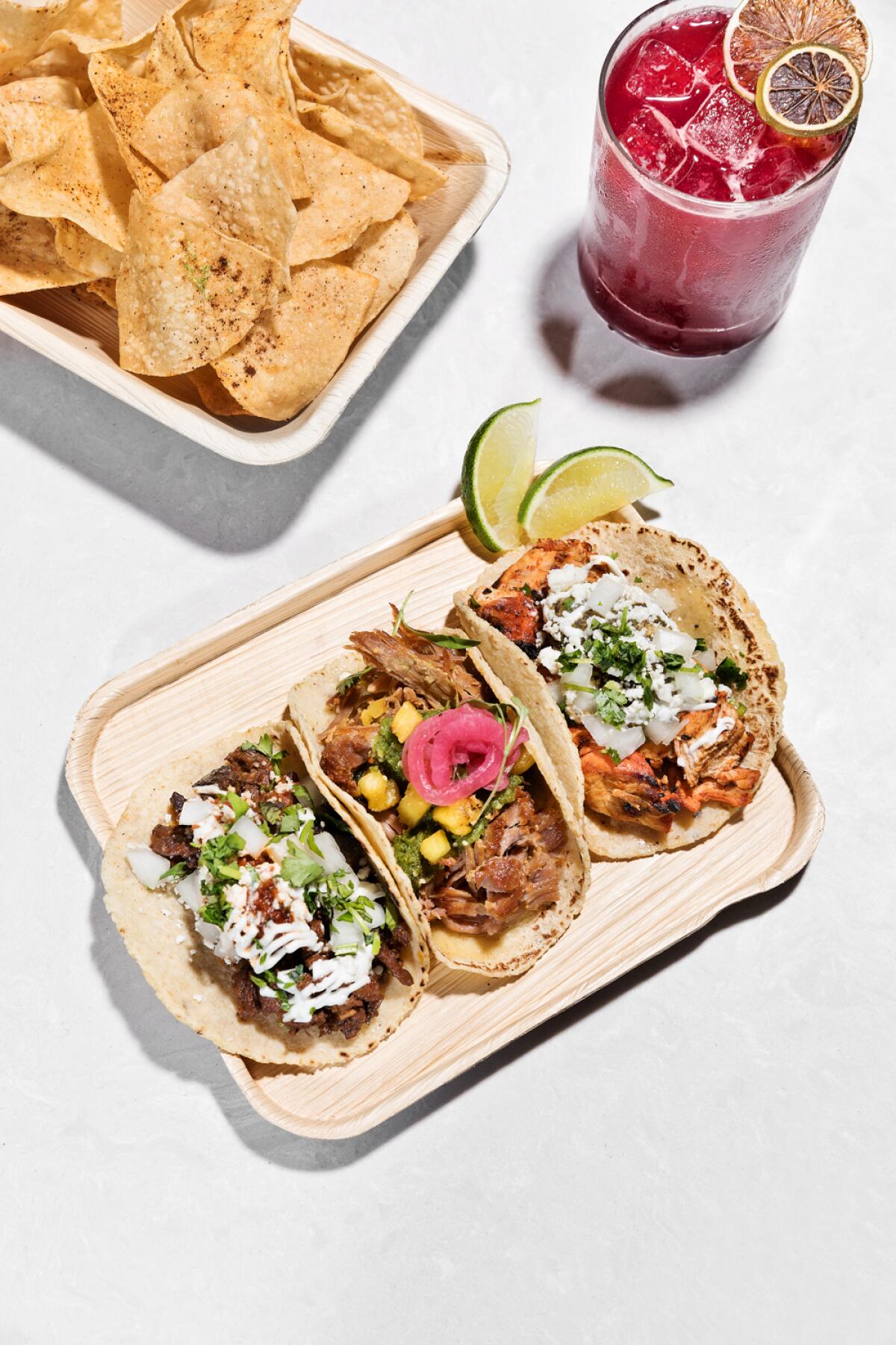 Tortilla chips and a trio of tacos with a pink beverage. 