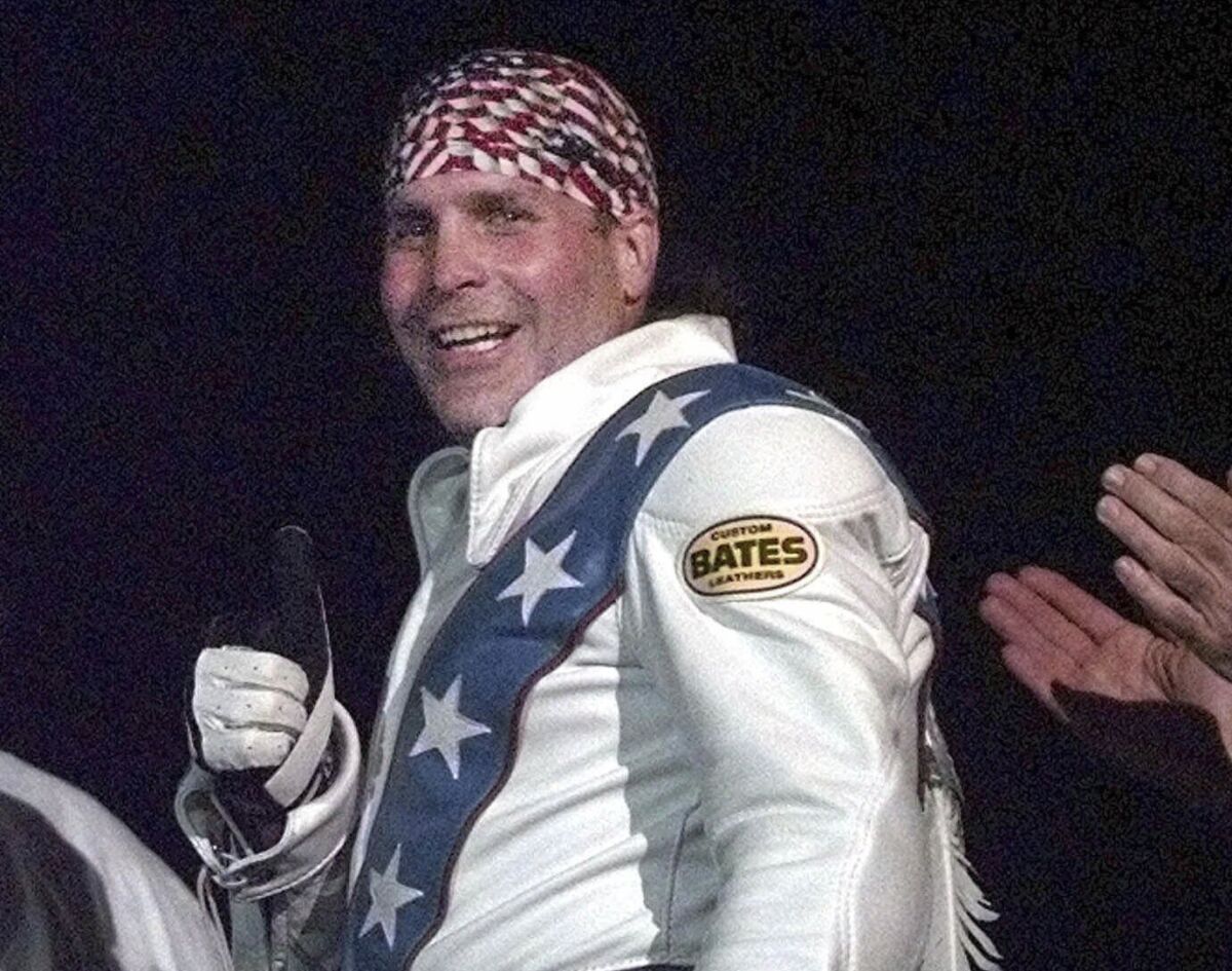 A man in an patterned bandana in a white jacket doing a thumbs up