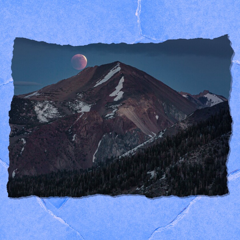 Shadow falls over a reddish moon during an eclipse. Below it is a mountain with scattered snow and green trees.