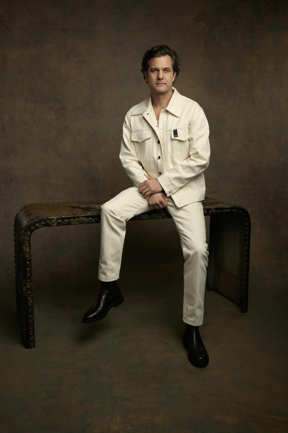 Joshua Jackson dressed in white sits on a table for a portrait.